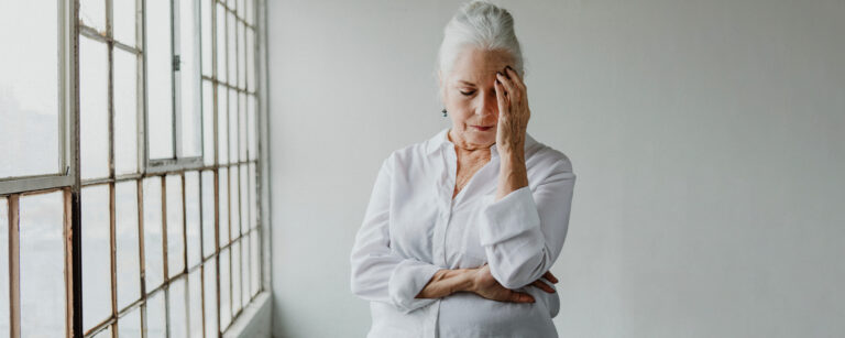 An older woman in a white shirt stands indoors by large windows, resting her head in one hand with a concerned or tired expression, possibly reflecting the lingering effects of head injuries.