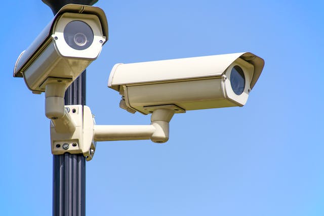 Two security cameras are mounted on a black pole against a clear blue sky, positioned to monitor different directions and capture comprehensive camera recordings.