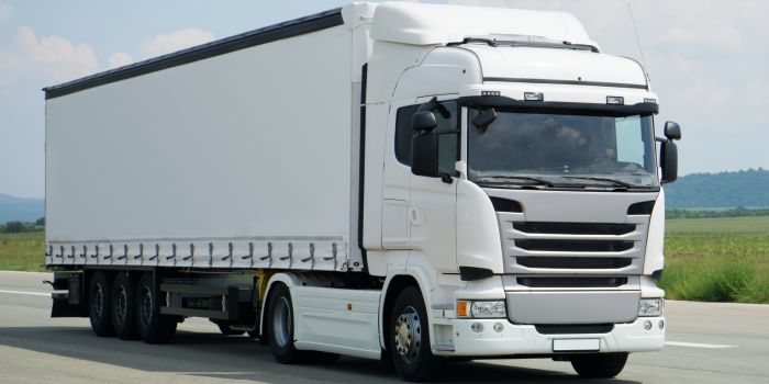 A large white semi-truck with a covered trailer is driving on a paved road with hills in the background.