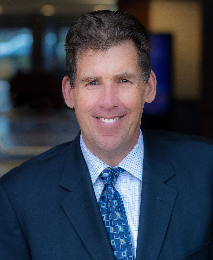 A man in a dark suit and patterned tie smiles at the camera with a blurred office background.