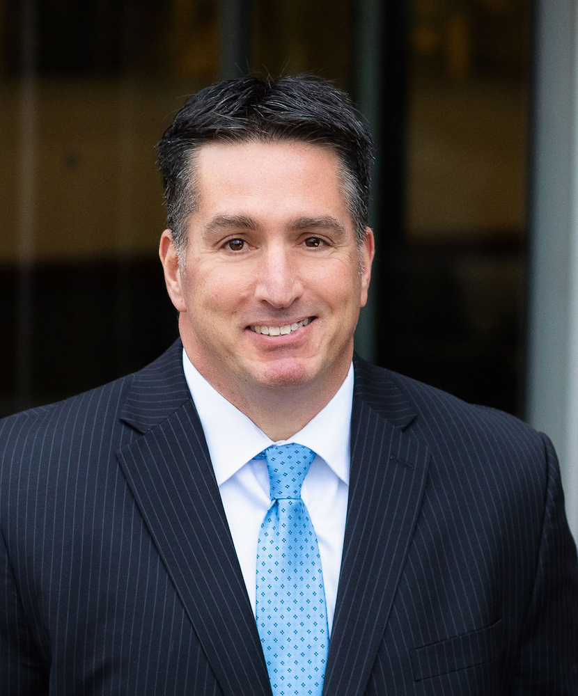 Man in a dark pinstripe suit and light blue tie, with short dark hair, standing in front of a blurred background outdoors, smiling at the camera.