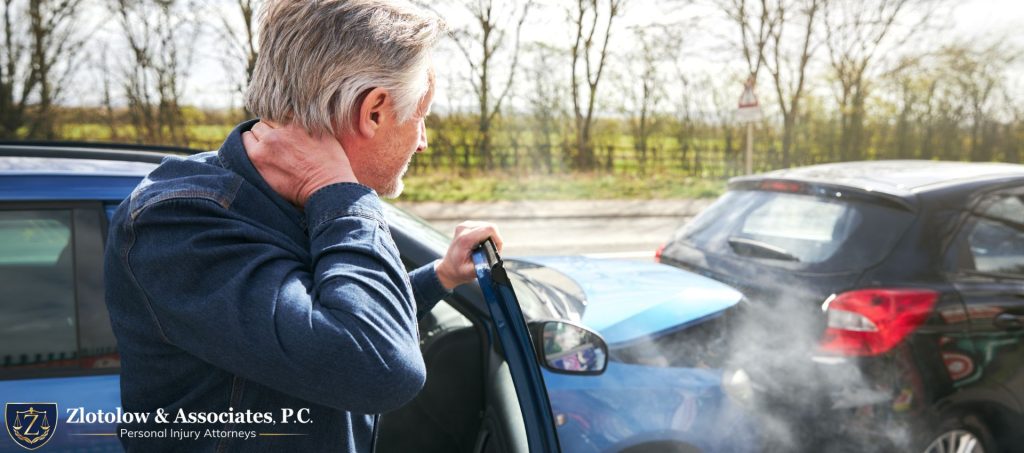 A man holding his neck stands next to a car with front-end damage after a collision, with another damaged car in the background.