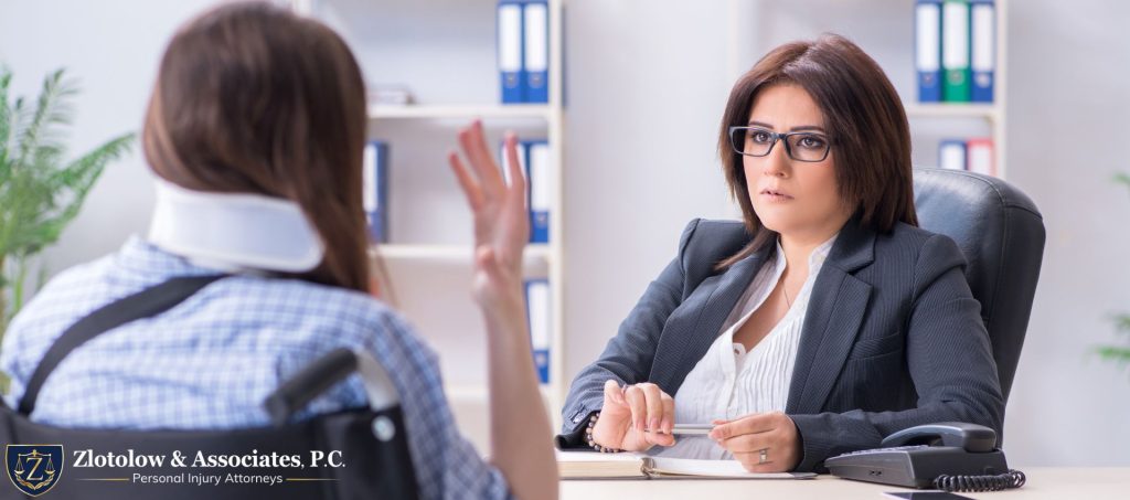 A woman with a neck brace speaks to a professional woman at a desk in an office setting. Logo for Zlotolow & Associates, P.C., Personal Injury Attorneys, is visible in the lower left corner.