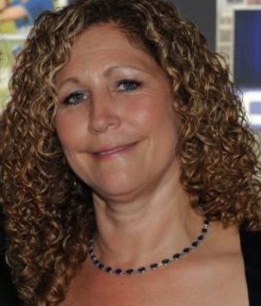A woman with curly blonde hair, wearing a necklace and a black top, smiles at the camera.