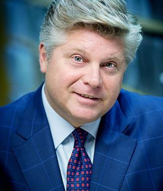 A man with light hair wearing a blue checkered suit and patterned tie looks toward the camera with a slight smile.